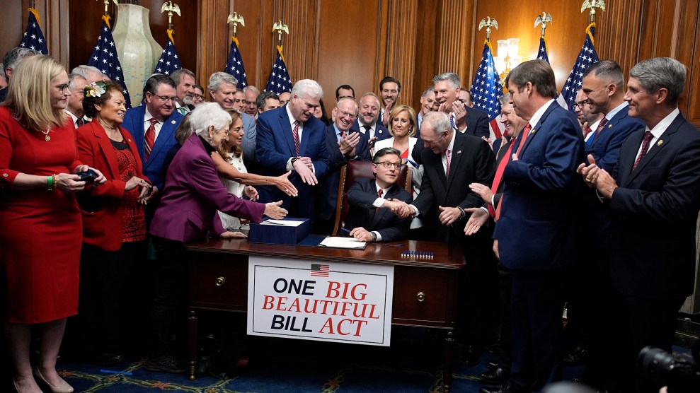 A group of over a dozen people dressed in suits or pantsuits. They clap and shake hands. Mike Johnson sits at a desk in the center of them. On the desk is a sign that says "One Big Beautiful Bill Act."