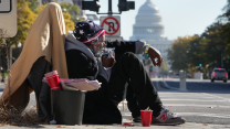 A man sits on a street corner amid a pile of his belongings on a sunny day. Smoke from a cigarette held to his mouth wafts past his face. In the background are trees lining a street and the dome of the US Capitol.
