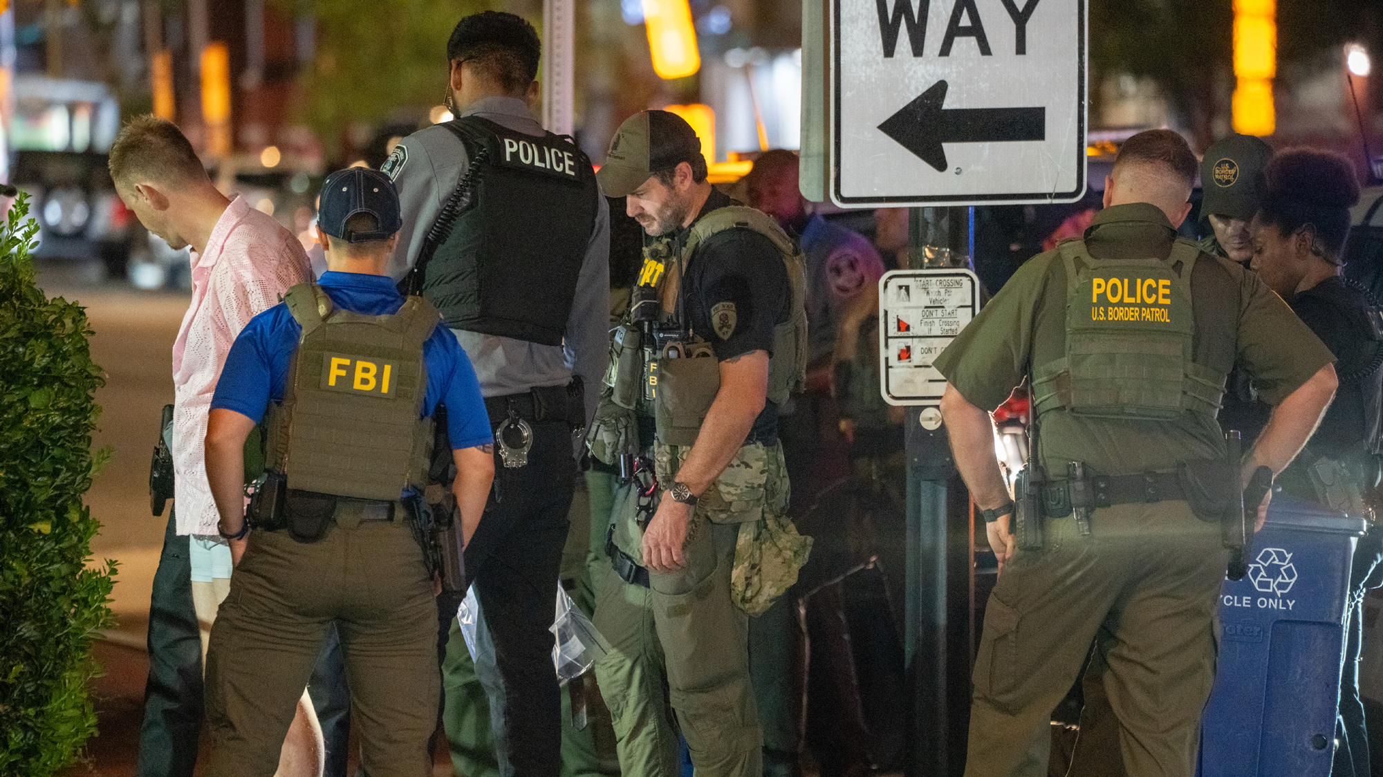 Officers in green uniforms, vests and baseball caps surround a man in a pink shirt and blue shorts.