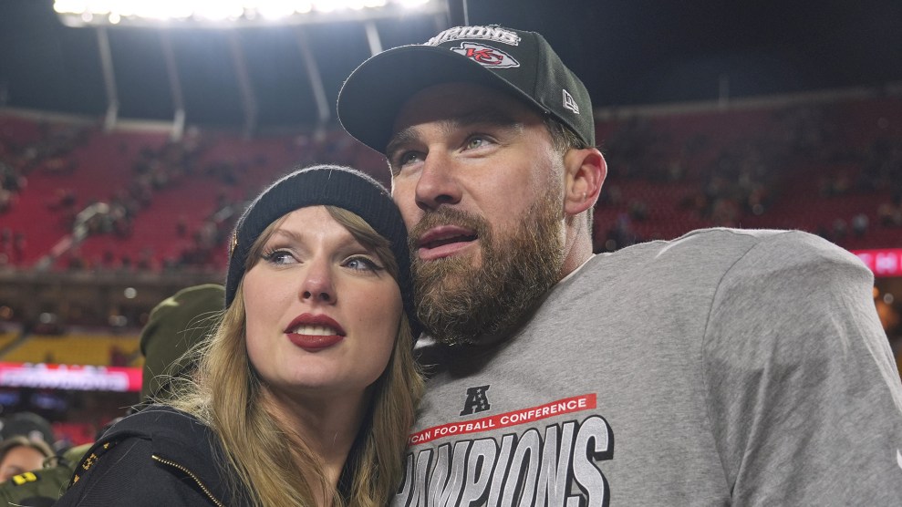 Taylor Swift and Travis Kelce pose for a photograph in a football stadium. She wears a black beanie and a black sweatshirt. He wears a Kansas City Chiefs cap and a grey t-shirt that says “Champions”