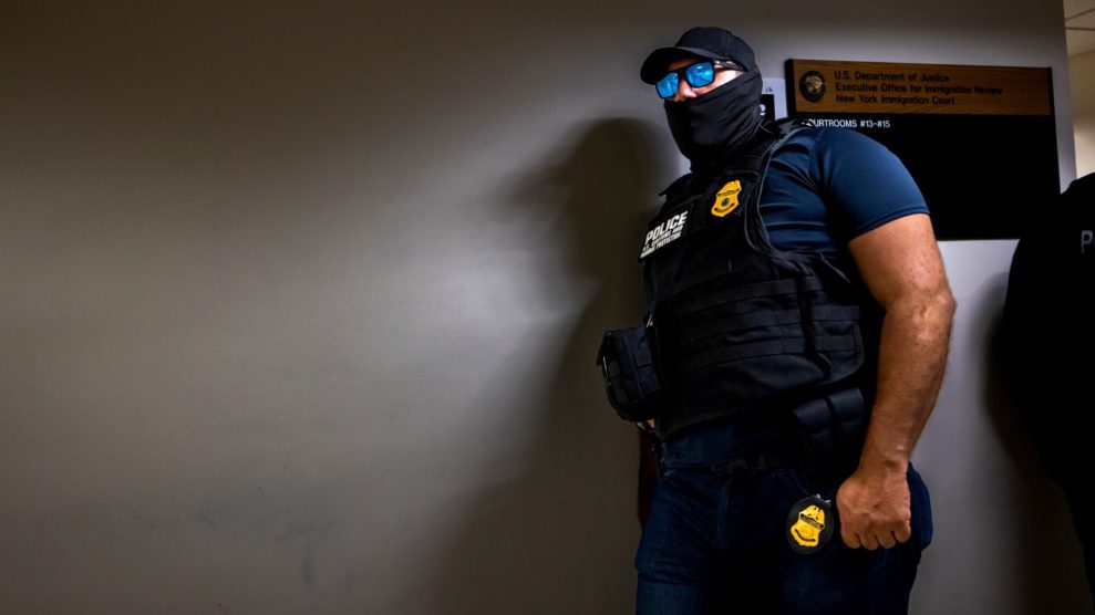 A masked ICE officer leans against a wall in a hallway waiting for a person to detain.