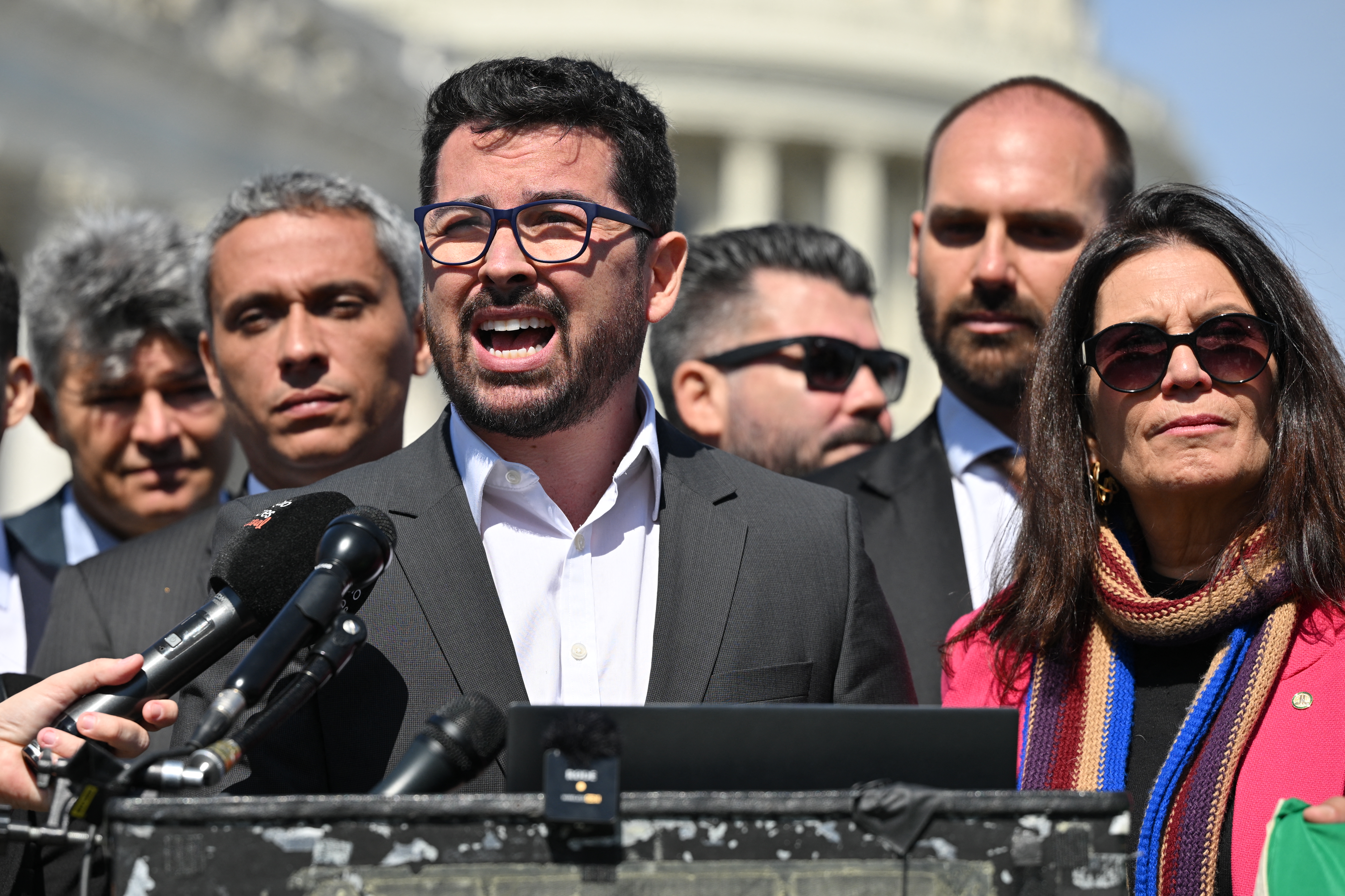 A bearded man in glasses and a suit speaking emphatically at a podium.