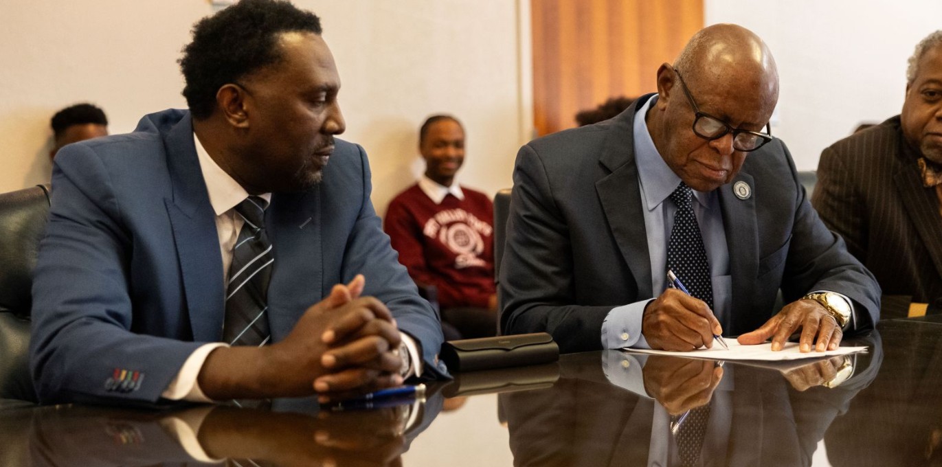 Two black men wearing suits sit next to each other at a wooden table. One signs a paper while the other looks at him.