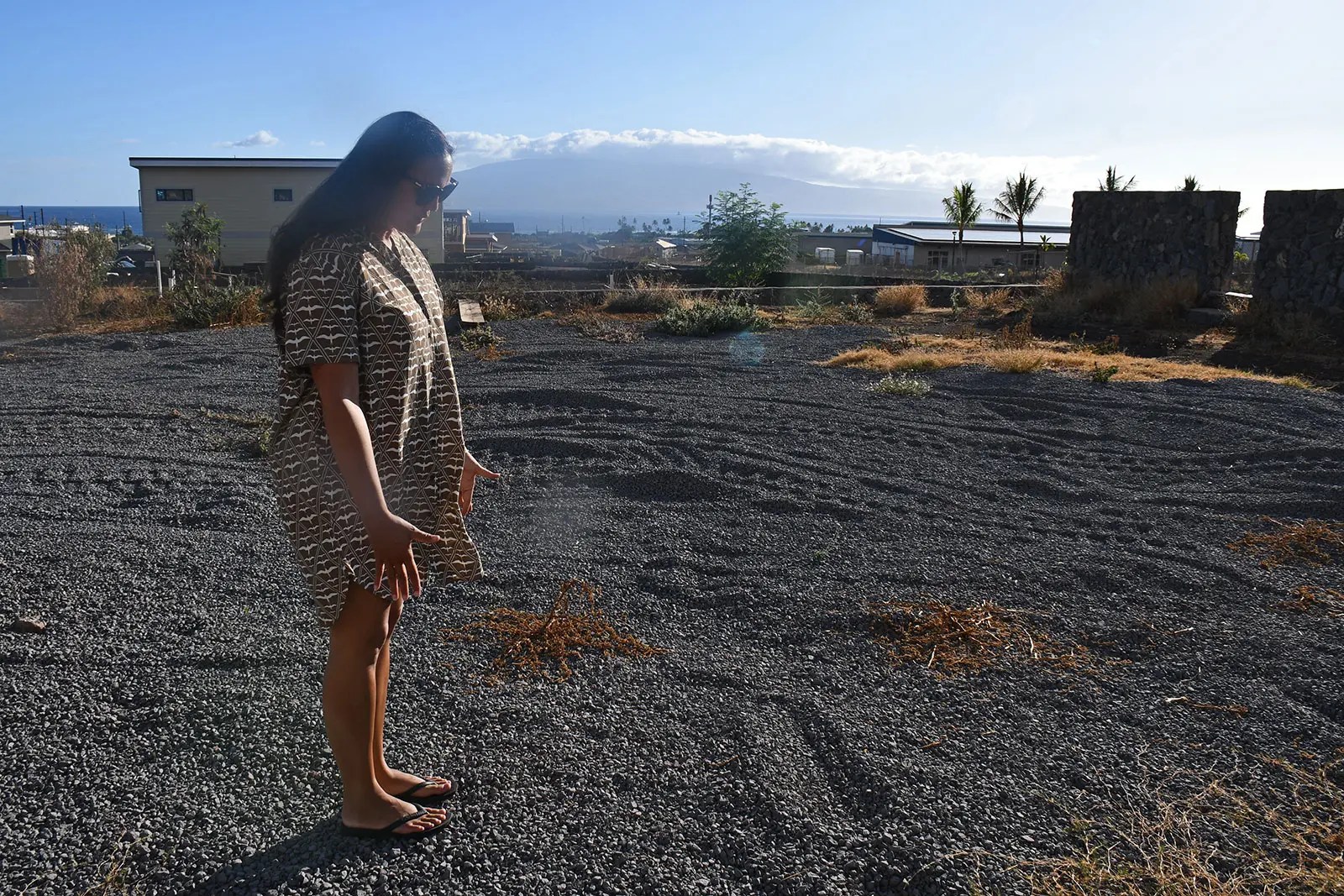 A woman wearing a dress looks down at black gravel.