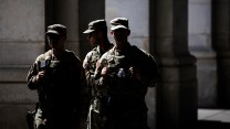 Three National Guard troops in camouflage uniforms stand in a cluster under the shade of a building. Their faces are obscured in shadow and in sharp contrast to the sunlight.