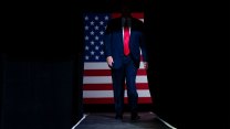 A shadow obscures the face of a man with a suit and red tie standing in front of an American flag.
