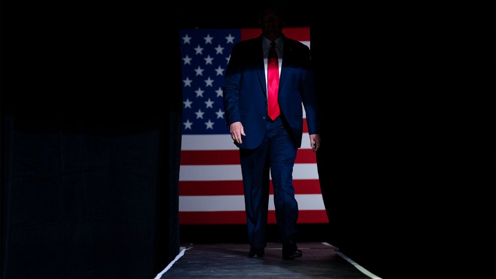 A shadow obscures the face of a man with a suit and red tie standing in front of an American flag.
