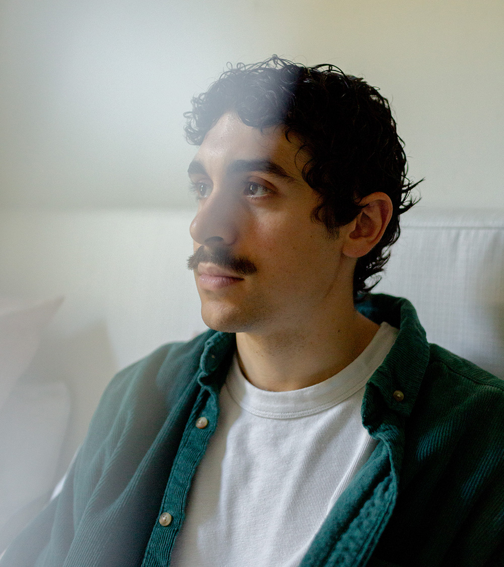 Portrait of young man with dark curly hair and a mustache. He wears a dark green button-up shirt open over a white T-shirt.