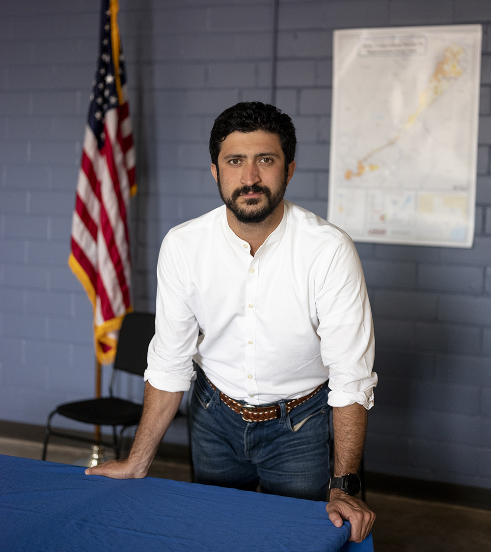 Greg Casar, with short dark hair and neatly trimmed beard and mustache, poses for a portrait wearing a white collarless button-down shirt and jeans. An American flag stands in the background.