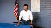Rep. Greg Casar, in a white button down shirt and jeans, poses with an American flag in the background.