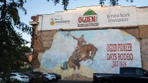 A mural on a brick building depicts a cowboy riding a bucking horse. Next to the cowboy, text reads: Ogden Pioneer Days Rodeo July 19-24.