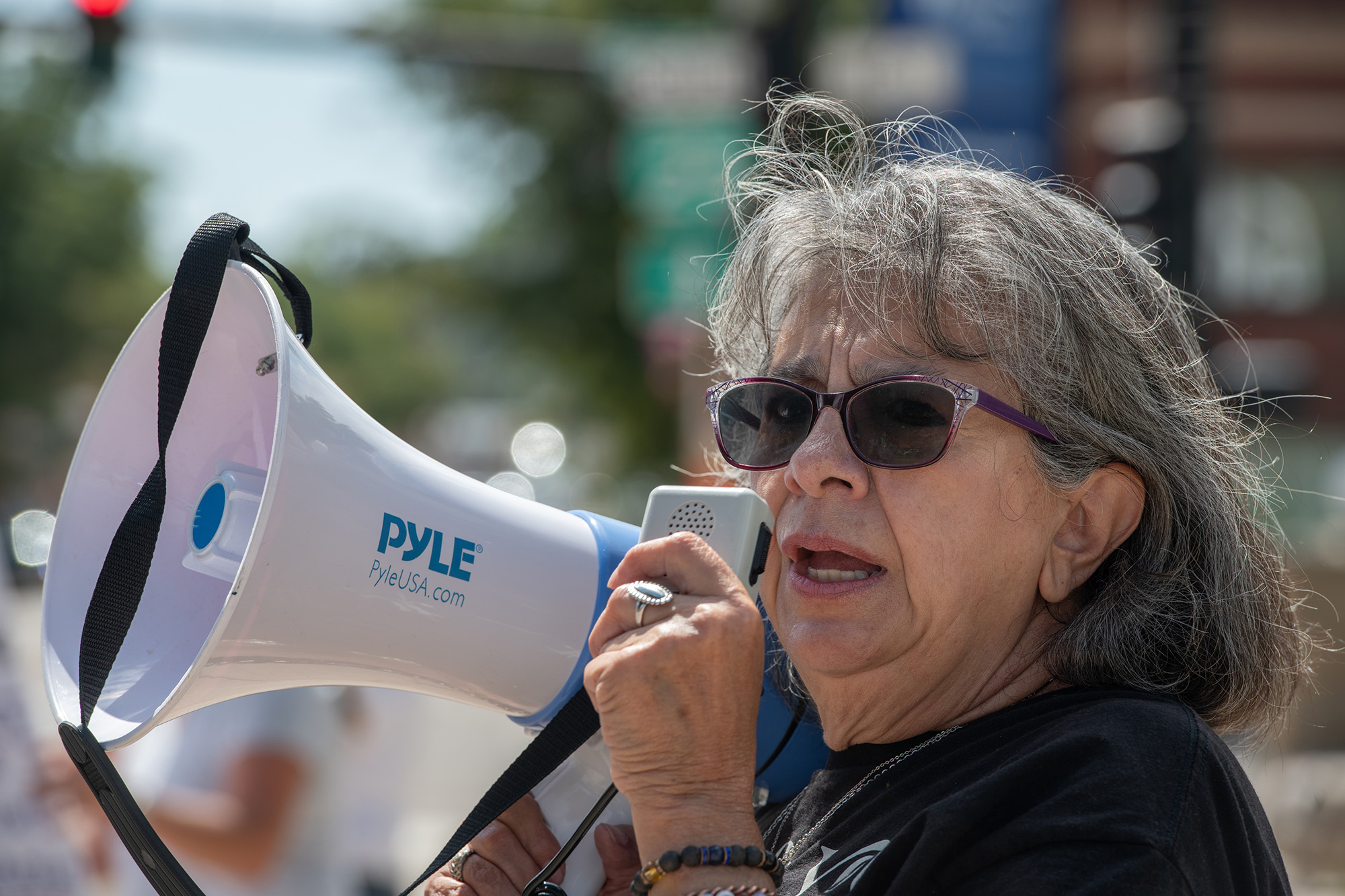 Dee Maes-Sandoval, wearing sunglasses, holds a speaker and  a megaphone to her mouth.