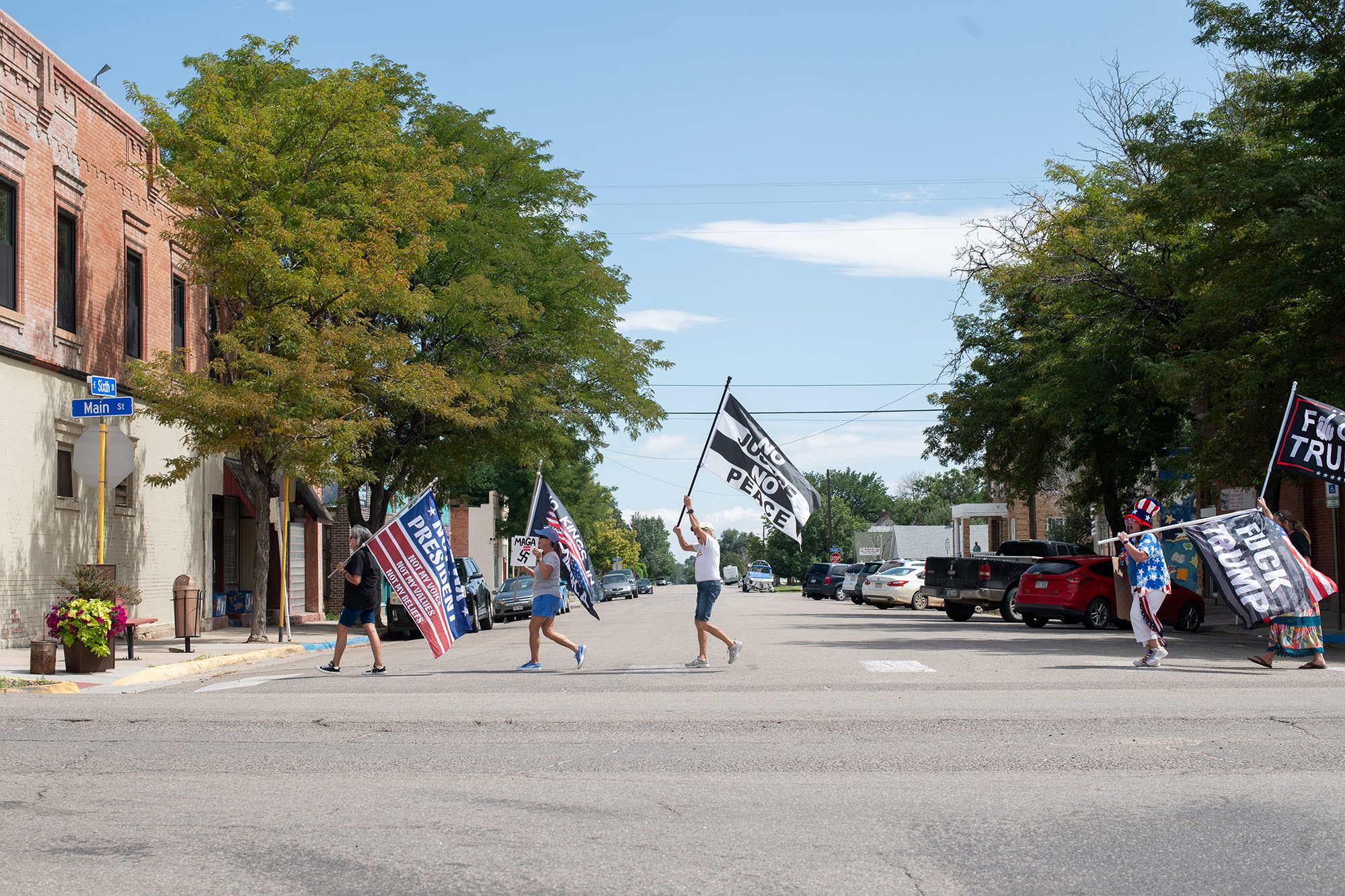 Protesters carry large flags as they cross a street on a sunny day. A black-and-white flag reads, "No justice, no peace." Two more flags read, "Fuck Trump."