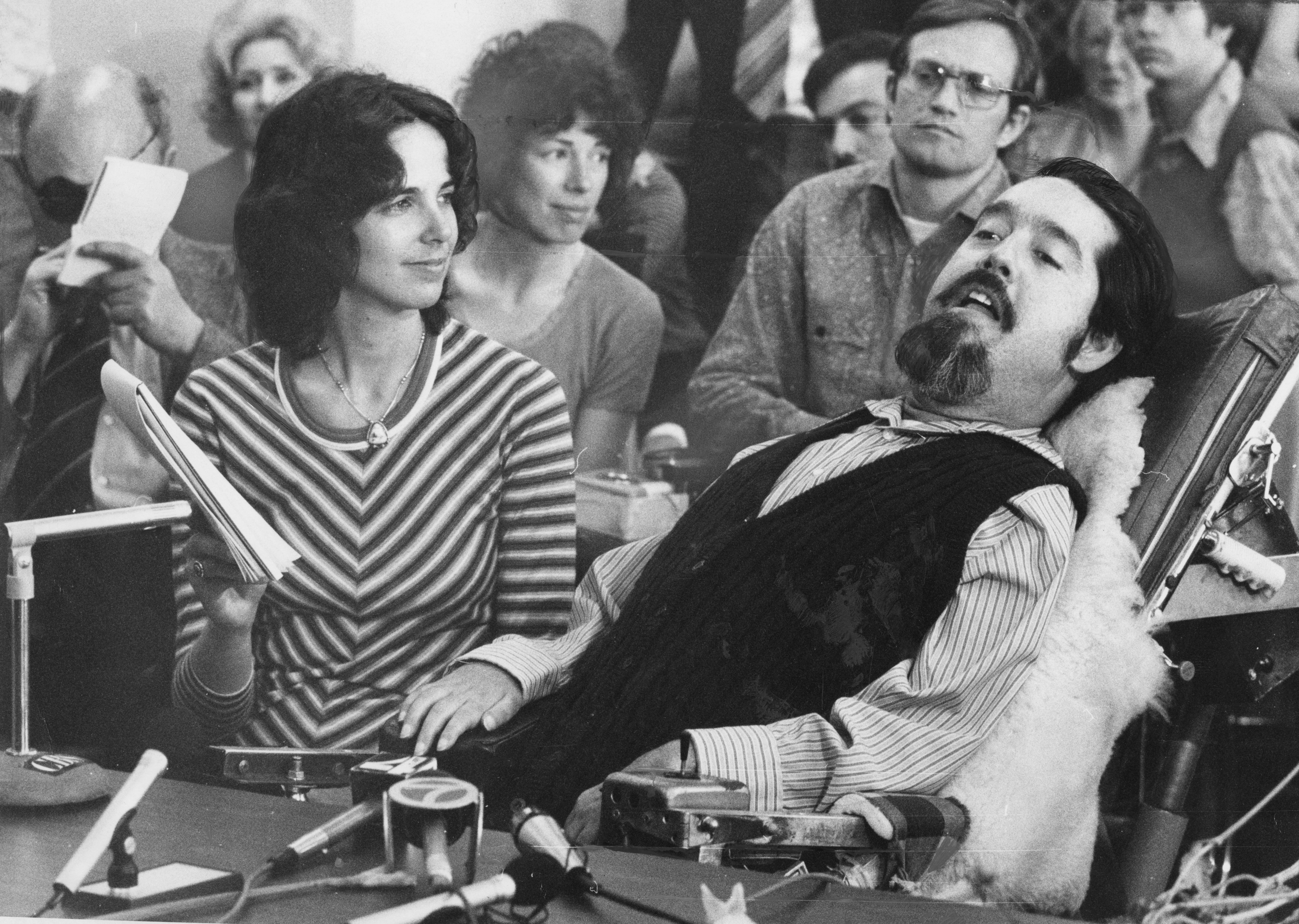 A man in a long-sleeve shirt and vest sits back in a mechanized chair, as a woman on the left holds up a pad. The man has dark black hair, a mustache and thick goatee. There is a gallery of people behind them.