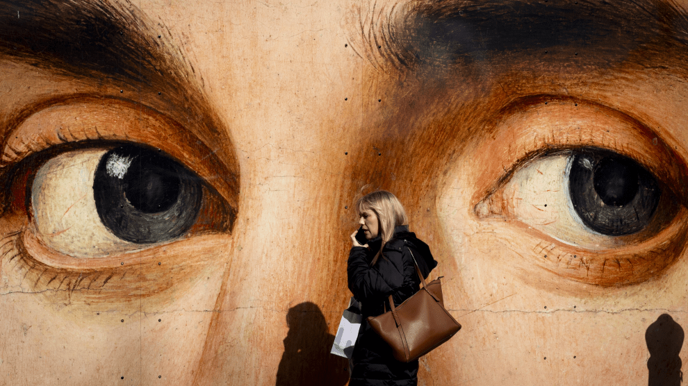 A Caucasian woman with hair down to her shoulders, wearing a coat and carrying two bags, walks to our left as she talks on a mobile phone. Behind her is a large mural of a face, seen from the bridge of the nose to the forehead. The dark gray eyes of the painting seem to be in line with the passing woman, whose shadow falls against the lower part of the wall.