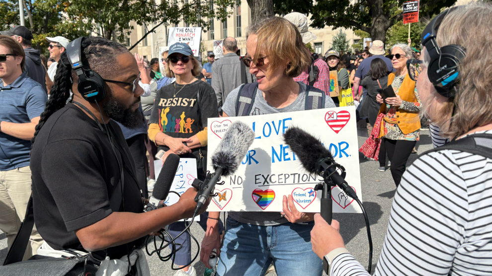 A man with long dreadlock hair and bushy beard holds a microphone in front of a woman with short brown hair. She is among a crowd of people wearing sunglasses, T-shirts, jeans and shorts, who are holding signs. The woman’s sign reads, “Love Your Neighbor. No Exceptions.”