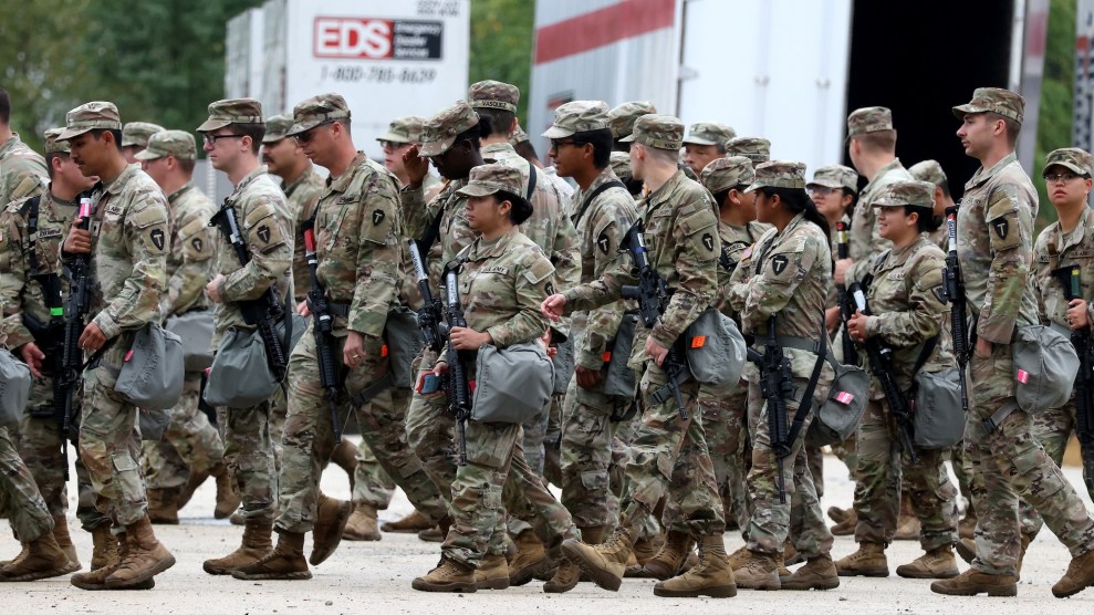 National guard officers in fatigues walk in loose formation in front of box trucks.