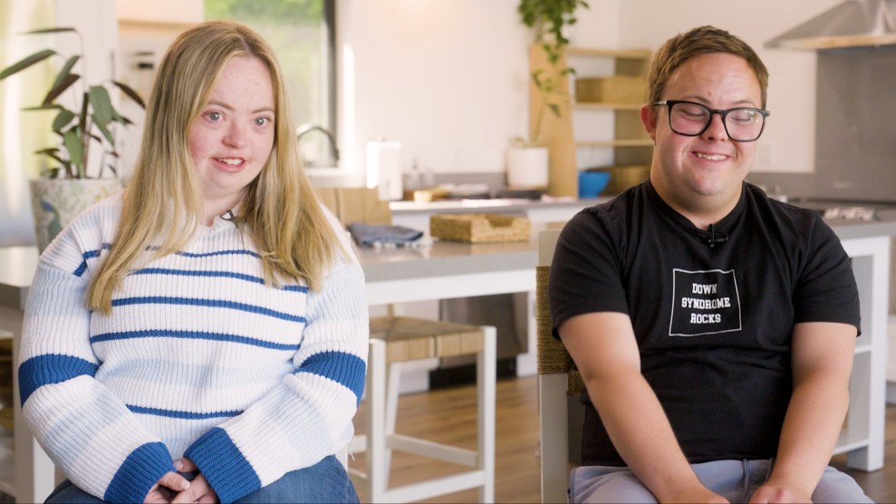 Two people sit in chairs in a kitchen facing the camera