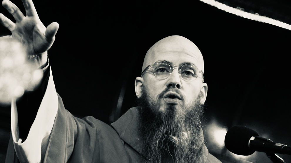 A black-and-white photo of a young Caucasian man with a shaved head, round wire-rimmed glasses, and long wispy beard. He wears unadorned liturgical vestments and is reaching out his right hand, as he appears to be speaking, as he stands behind a microphone.