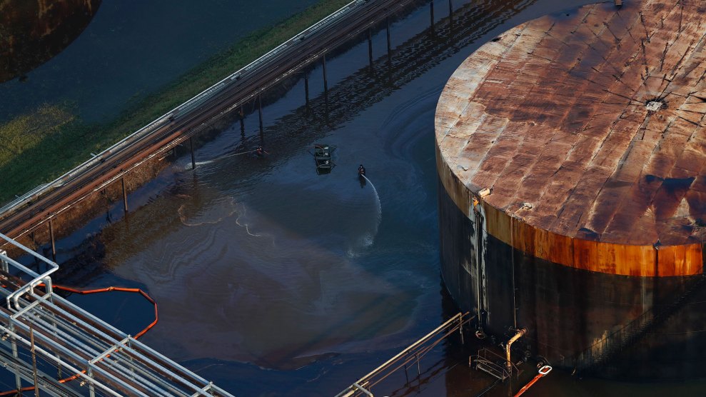 A worker does cleanup in shallow floodwaters next to a giant, rusting oil storage tank.