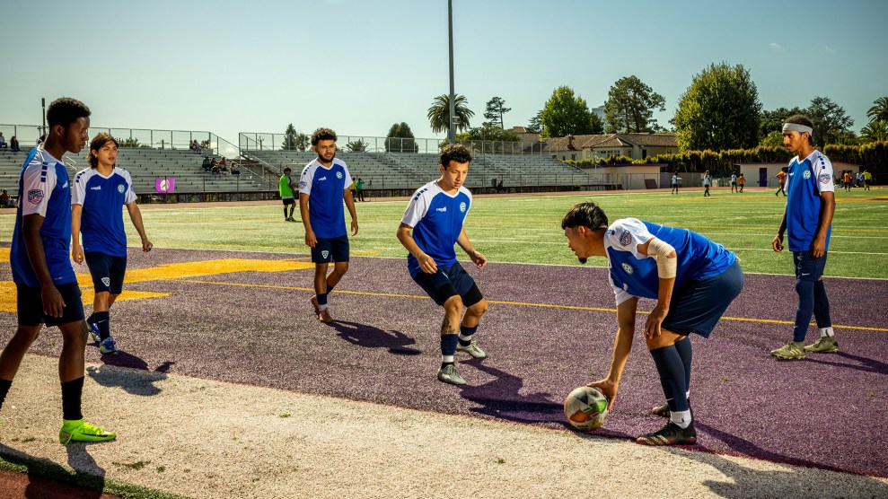 Six young men in blue and white jerseys practice on an astroturfed soccer field on a sunny day.
