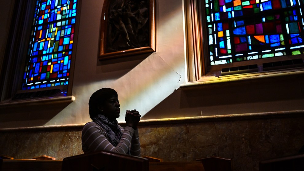 A person seen in silhouette kneels with hands clasped in front of their face. Behind the person are two tall stained-glass windows, made up mostly of rectangles in shades of blue, illuminated by sunlight.