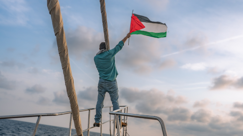 A man, seen from behind, stands on the railing at the bow of a sailboat. He wears a turquoise long-sleeve shirt and blue jeans, with a keffiyeh around his neck. He holds a Palestinian flag in his outstretched right hand. Around him, the water is dark and choppy and the sky dotted with gray clouds.