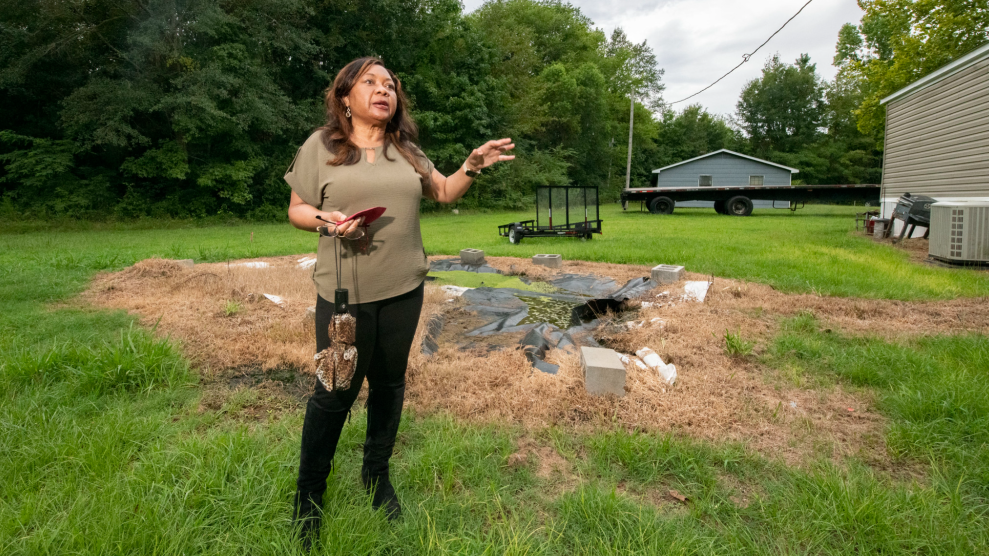 An African American woman with long hair, wearing an olive green blouse and black jeans, stands in a yard, steps in front of an area where the grass is muddy and brown, with a black tarp haphazardly covering some spots.