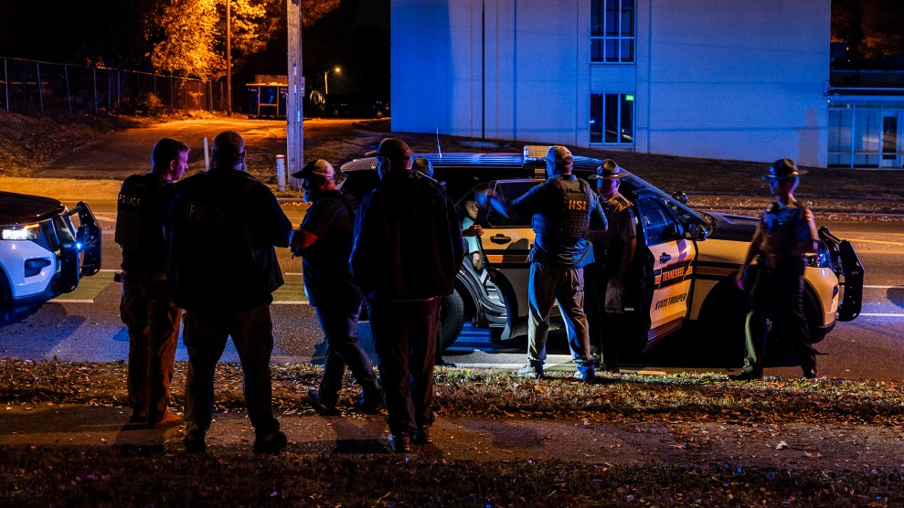 A group of plainclothes federal agents and uniformed state troopers stand around a stopped Tennessee State Troopers car at night. A building in the background glows blue from the reflection of nearby police lights.