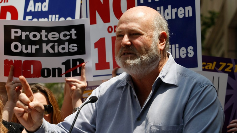 Rob Reiner speaking into a microphone while activists hold up signs at a rally.