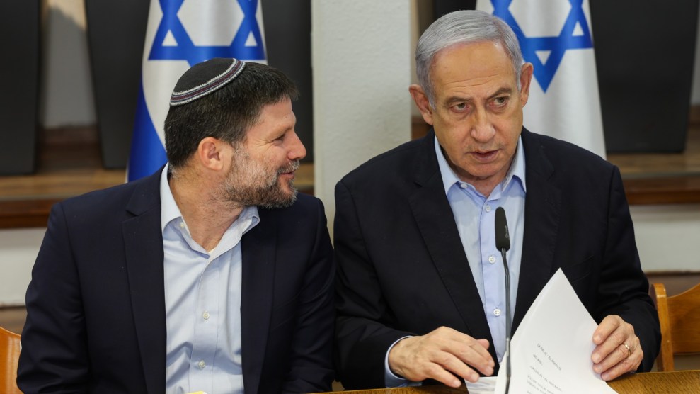 Two men site at a table. Man on the left looks at man on the right, who is facing forward clipping through papers. Two Israeli flags are in the background.