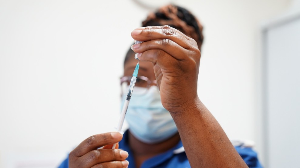 A nurse with a blue shirt fills a syringe with a Covid-19 vaccine dose. The nurse is holding a vial with the dose in their left hand and the syringe in their right.