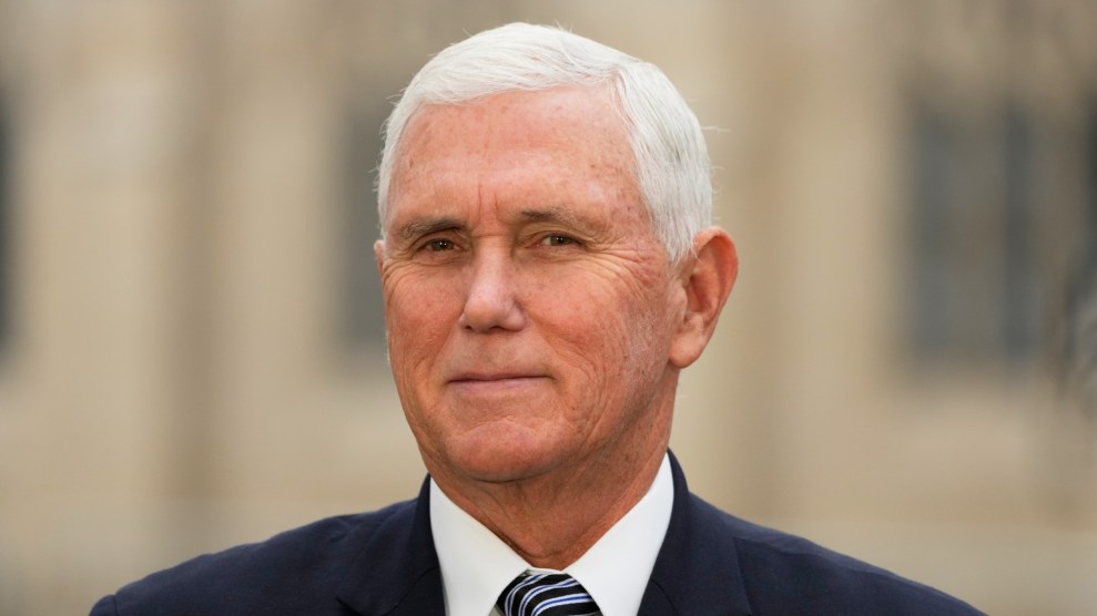 Mike Pence is shown facing forward. He is wearing a suit and a blue striped tie. The background is a building that is blurred out.