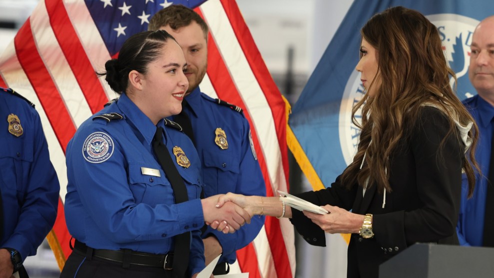 Kristi Noem shaking hands with a TSA officer dressed in a uniform. The American flag and other TSA officers are in the background.