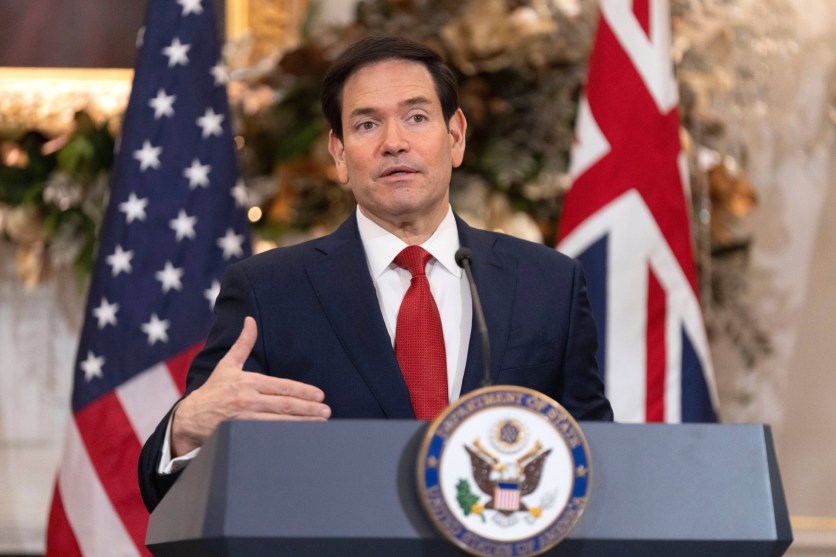 Marco Rubio dressed in a suit with a red tie stands behind a podium and gives a speech. The USA and Great Britain flags are in the background.