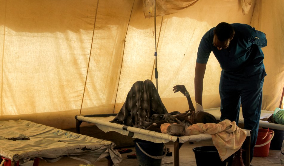 A man leans down to a bed in a tent and touches a man laying down on the bed, sick.