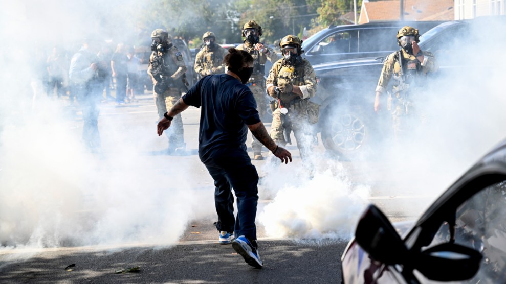 One person in the foreground stands facing away from the camera while five federal agents stand in the background with gas masks. Tear gas has been thrown on the street in a protest.