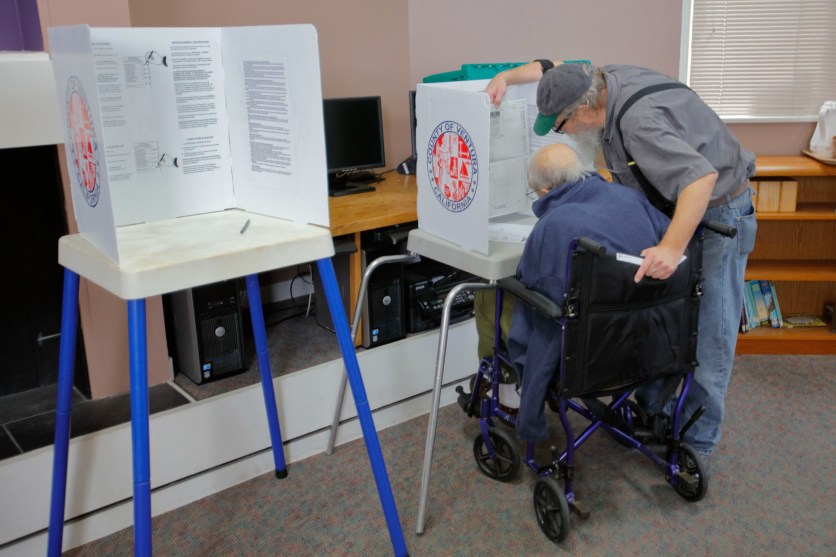 Senior man helps a man in a wheelchair vote at polling station.