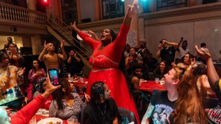An African American woman in a formal, bedazzled red dress sings with her arms raised among people seated at long tables draped with red tablecloths.