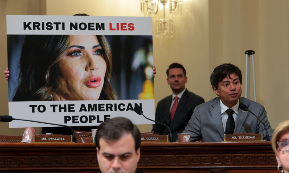 Shri Thanedar, a South Asian man with black hair in a gray suit and black tie, sites next to a poster with an image of Kristi Noem and the text "Kristi Noem Lies to the American People."