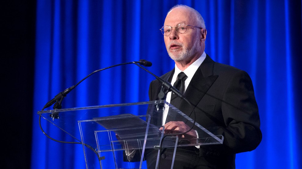 A white haired white man with a beard wearing a black formal suit, stands at a plexiglas podium with blue-lit curtain behind him