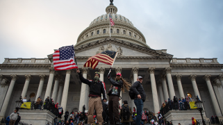 Three men stand atop a vehicle in front of a crowd of others outside the US Capitol. They are dressed in MAGA gear and wave American flags. Many in the crowd wear red MAGA hats.
