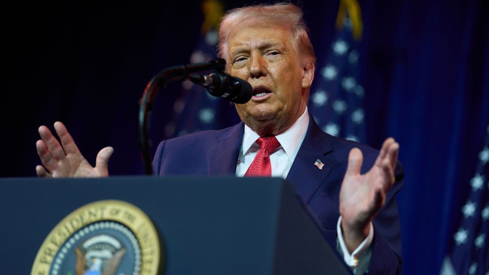 A photograph of Donald Trump wearing a blue suit and red tie standing behind a podium, speaking into a microphone.