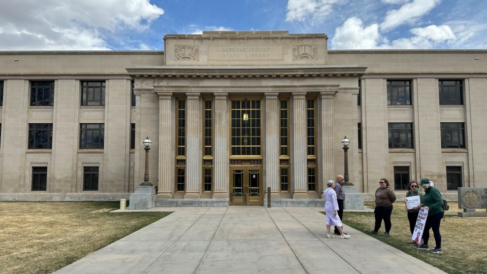 A view of the front of the Wyoming Supreme Court building. The sign on the building reads: "SUPREME COURT STATE LIBRARY." Five people stand in front of the building. Two of them are holding signs. One of the signs mention "birth control."