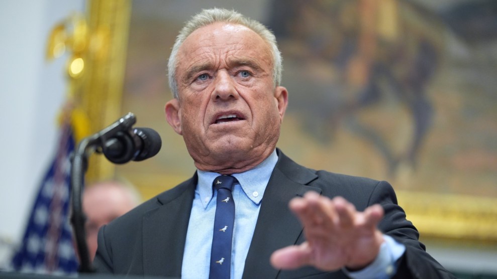 Robert F. Kennedy, Jr., speaks into a microphone on a podiium. He is wearing a black suit with a blue tie. A painting and the American flag are shown blurry in the background.