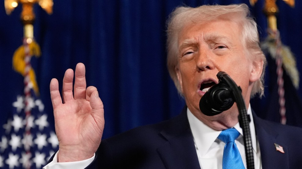 President Trump dressed in a black suit with a blue tie and American flag pin is speaking into a microphone at a press conference. He is holding up his right hand with thumb and second finger touching.