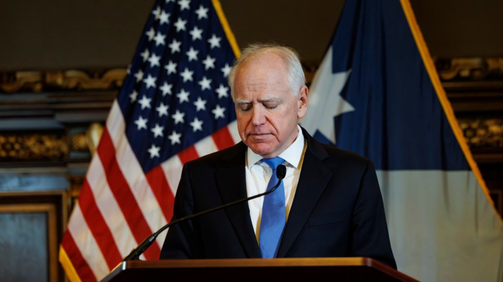 Governor Tim Walz is wearing a black suit and blue tie. He is standing at a podium with a microphone and looking down. The American flag is in the background.
