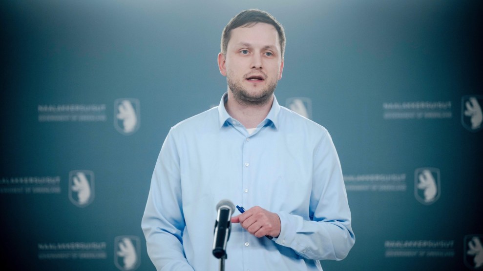 Greenland Prime Minister Jens-Frederik Nielsen speaks at a press conference. He is standing at a podium with a microphone. He is wearing a blue dress shirt and carrying a pen in his left hand.