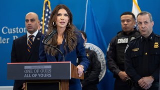 Kristi Noem stands at a podium with a microphone. She is wearing a blue suit and her hands are placed on the podium. Four people are standing in the background behind Noem. The podium lists the DHS website and has a message reading, "ONE OF OURS ALL OF YOURS."