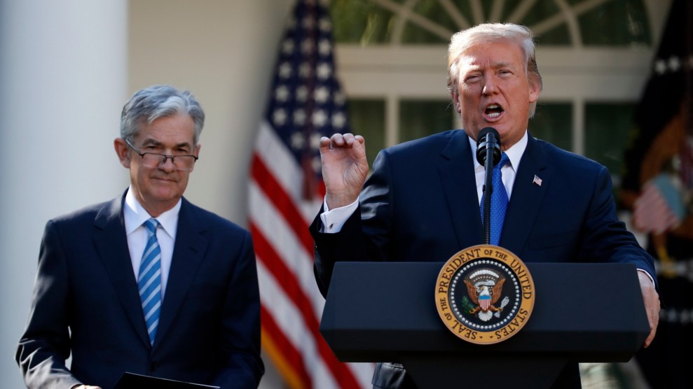 President Trump stands on the right on a podium and speaks into a microphone. He is wearing a black suit and a blue tie. His left hand is on the podium and his left hand is making a circle to emphasize his speech. Jerome Powell, stands behind and to the left. He is wearing a black suit and a stripped blue tie.